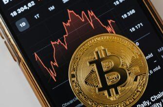 A close-up of a gold Bitcoin coin placed on a smartphone displaying a stock market chart.